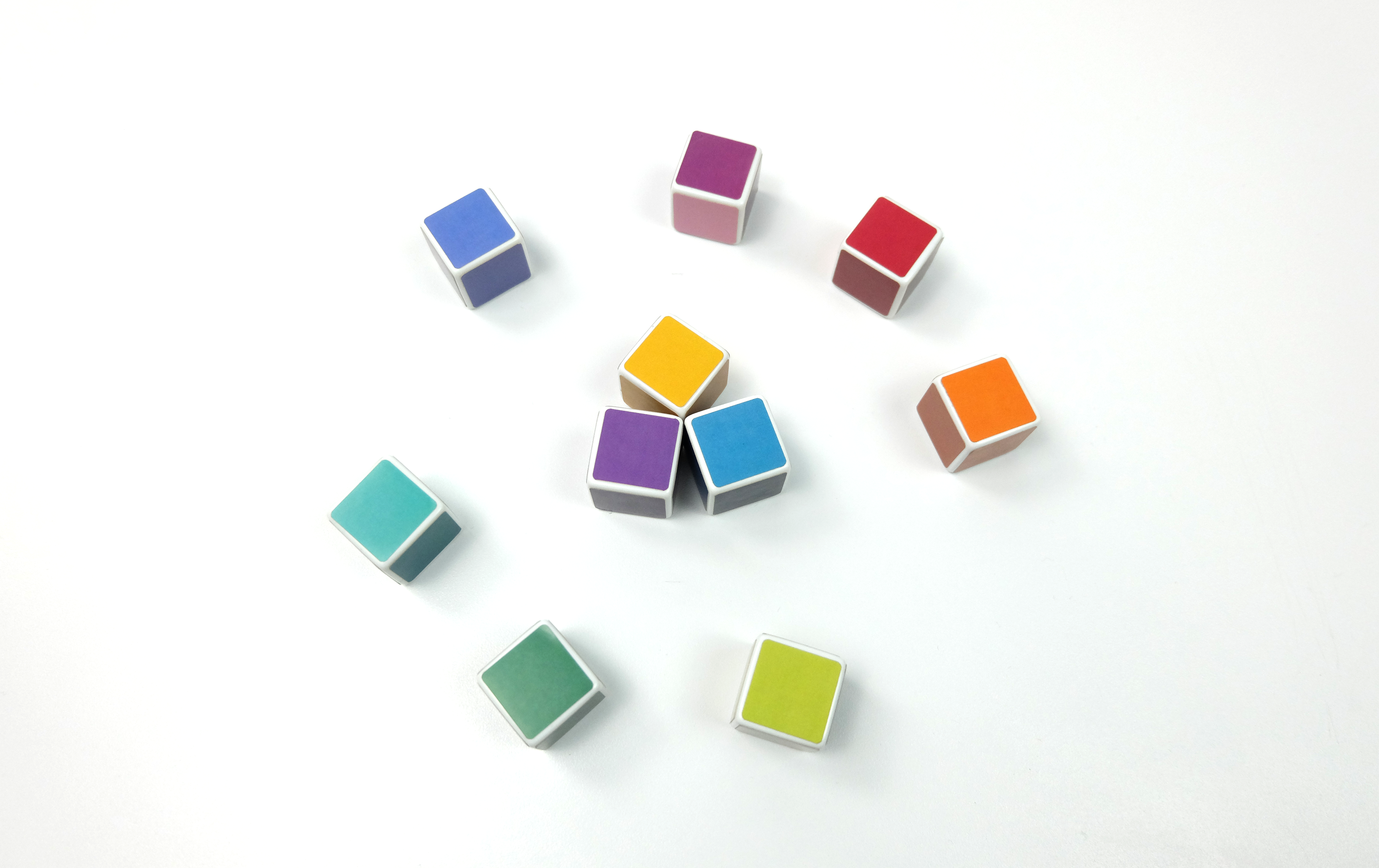 Three colorful dice are displayed together inside of a larger ring of colorful dice arranged in a color wheel on a white table.